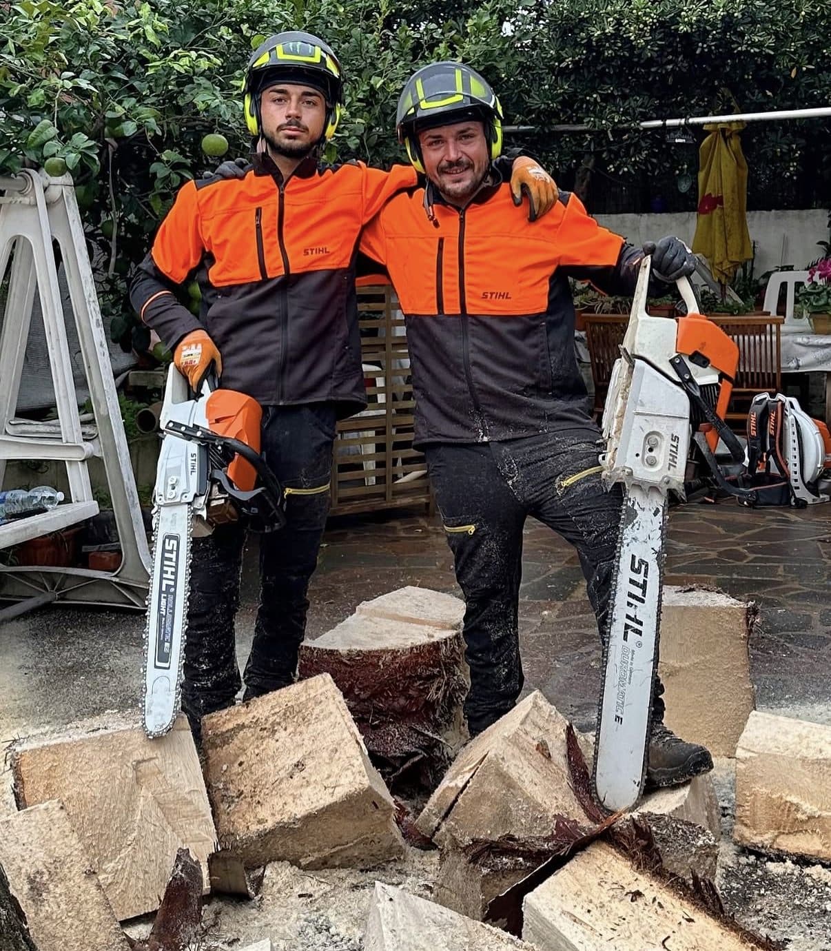 Gianluca e Federico in giacca antitaglio e casco con visiera, ognuno con la sua motosega, davanti al tronco appena abbattuto e sezionato in ceppi