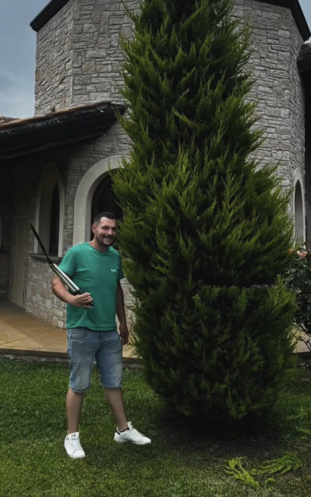 Operatore di GF Garden con il tagliasiepi ad asta accanto a un cipresso appena sagomato, alto quanto la casa alle sue spalle