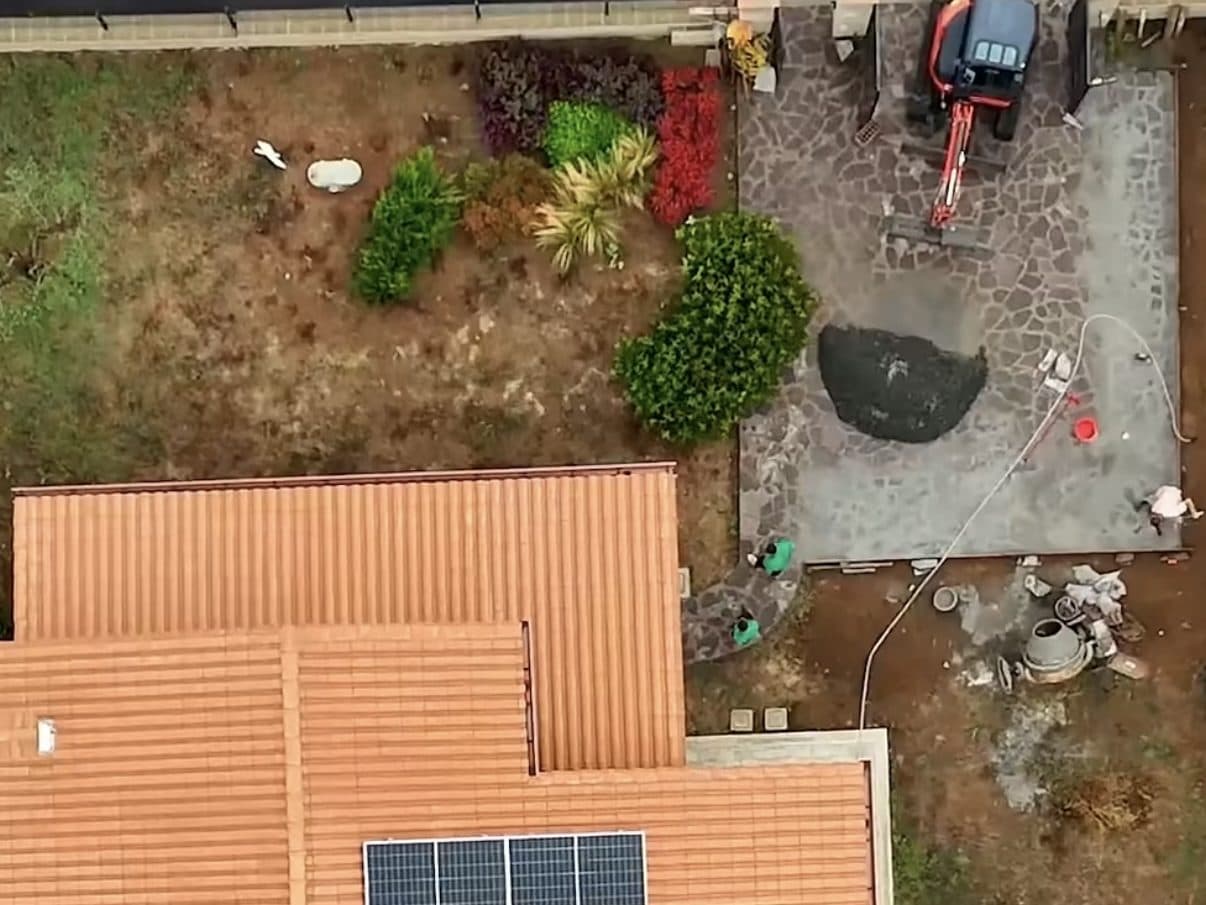 Il giardino durante i lavori, visto dall’alto: terreno ancora spoglio, betoniera, miniescavatore e la pavimentazione in pietra in corso di posa