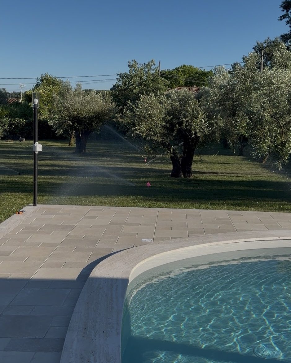Irrigatori in funzione fra gli ulivi al tramonto, con il getto che attraversa il prato accanto al bordo della piscina